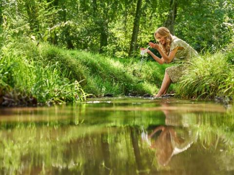 Eine Frau sitzt am Ufer eines Baches im Wald. Mit den Händen schöpft sie Wasser.